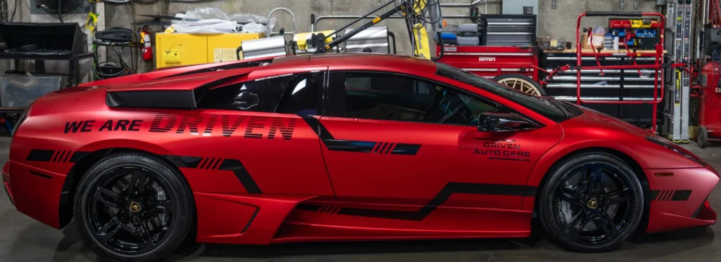 Matte red Lamborghini wrapped with black graphics parked inside a professional auto shop. Large side lettering reads We Are Driven and the door displays Driven Auto Care, highlighting a custom commercial vehicle wrap with bold branding and performance styling.