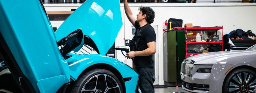 Technician inside an auto detailing shop lifts the hood of a teal supercar while holding a heat gun during paint protection film installation. The image shows a professional applying clear bra PPF with precision tools to ensure smooth adhesion and full surface coverage. This highlights expert installation techniques used to protect high end vehicles from chips and surface damage.