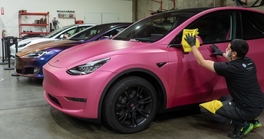 Matte pink Tesla Model Y inside a custom shop with a technician working near the rear door. A dark overlay reads Tesla and EV Customization with a View Service button, showcasing electric vehicle wrap and customization services.