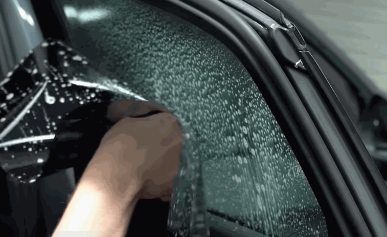 A technician installs wet window tint on the side window of a dark vehicle. Water droplets cover the glass as the film is positioned for a smooth finish.