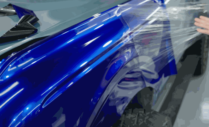 Glossy blue color change vinyl wrap is applied across the rear fender of a vehicle. A hand smooths the clear backing as the film conforms around the wheel arch.