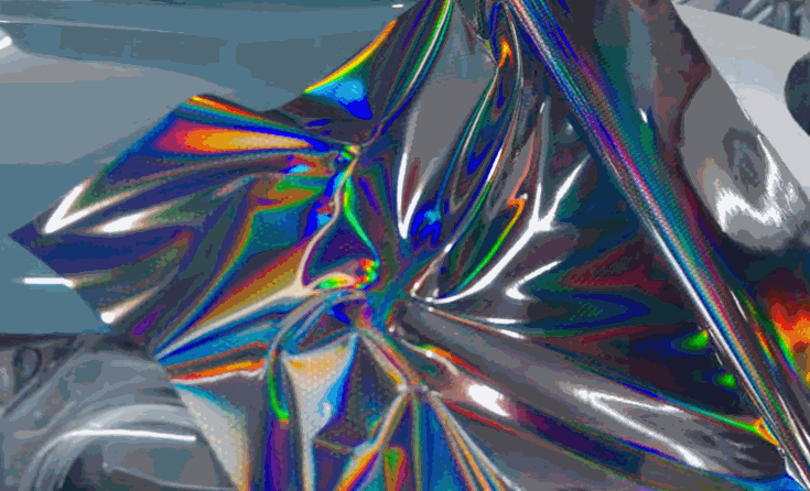 Iridescent color change vinyl wrap is stretched over a vehicle panel with rainbow reflections across the glossy film.