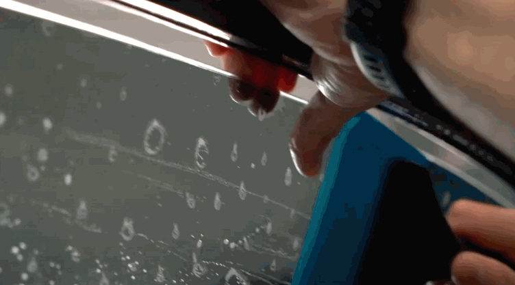 A technician trims wet window tint along the top edge of a car window. A blue squeegee holds the film steady against the glass.