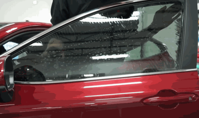 Window tint covers the front side window of a red vehicle during installation. Water droplets show the film being positioned for a smooth fit.