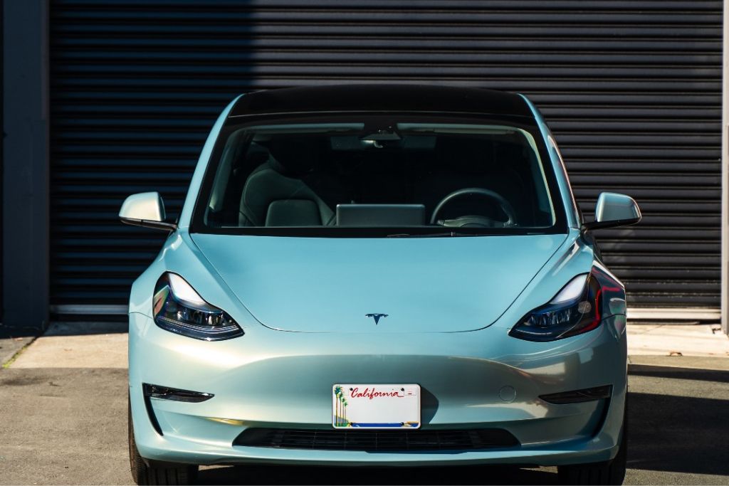 A light blue Tesla is parked front facing outside a shop with a dark roll up door behind it. The California license plate reads "California" and the clean front bumper and hood show areas where Tesla PPF can help protect against freeway debris and chips.